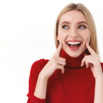 Beauty, women and fashion concept. Close-up portrait of silly adorable young blond woman in red turtleneck, smiling happy, poke cheeks and looking away at promo banner, white background
